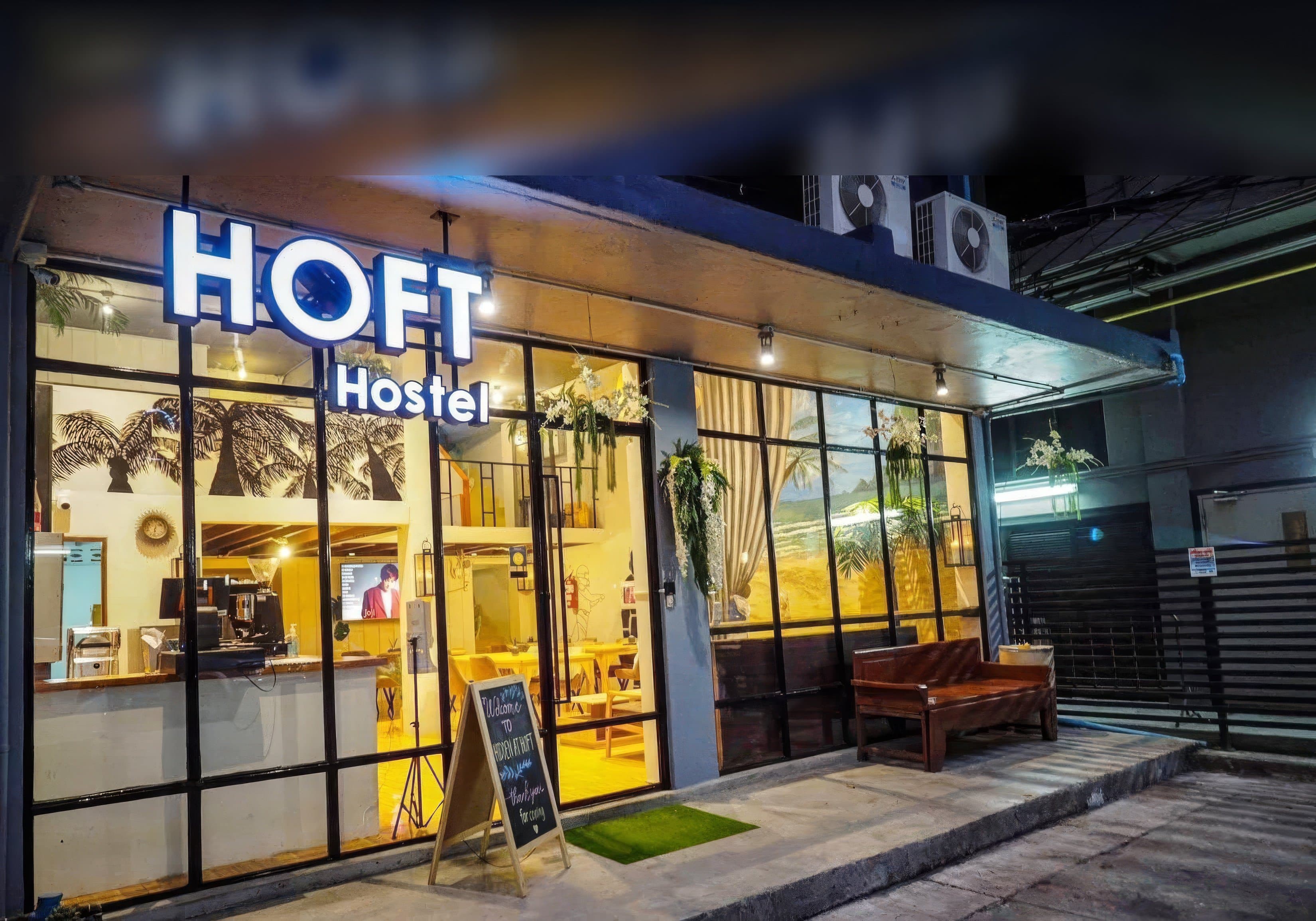 The lit HOFT Hostel sign above the glass frontage of Arawana Hoft Hostel in Silom, Bangkok at night, with a wooden bench on the pavement and the cafe and reception visible inside.